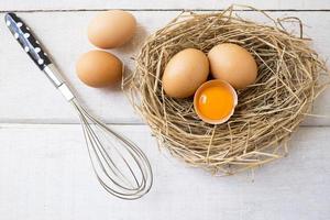 Fresh organic eggs on nest with whisk on white wooden background. photo