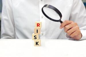 Hand of businessman holding glass magnifying over wooden blocks tower with text RISK. Concept of risk analysis, business management and strategy. photo