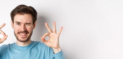 cerca arriba de hermoso barbado hombre parpadeo y sonriente, demostración bueno señales, asegurar todas bien, en pie en blanco antecedentes foto