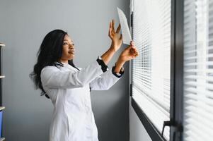 Young attractive african female doctor radiologist looking at x-ray image, while standing in light modern office in clinic photo