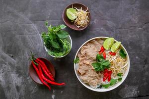 un cuenco pho bo tradicional sopa con carne de res, arroz fideos, jengibre, Lima, chile pimienta en bol. cerca arriba. vietnamita cocina servido en negro antecedentes foto