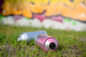 Several used spray cans with paint and caps for spraying paint under pressure on grass near the painted wall in colored graffiti photo