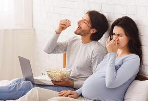 Unpleasant symptoms of pregnancy concept. Pregnant woman closing her nose, feeling sick with nausea because of popcorn that her husband eating at home photo