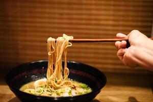 Crop anonymous female with chopsticks eating yummy ramen soup with noodles and meat in black bowl at ramen restaurant. photo