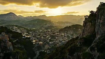 Serene aerial view of a mountain village bathed in golden sunset light, surrounded by lush hills and rocky cliffs under a dramatic evening sky. video