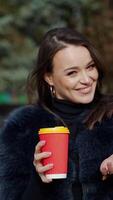Portrait of a happy girl in a sunny day in winter. Beautiful woman in fur coat holds a cup of hot drink in the park and smiling at camera. Slow motion. Vertical video