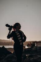 The silhouette of a female photographer confidently holding a camera on her shoulder stands out against the backdrop of a serene sunset. Her long ponytail and stylish scarf add a touch of creativity photo