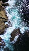 Aerial view. Ocean waves crash against rocky cliffs, forming white foam. A top-down view of a rugged coastline with a slow movement up and away video