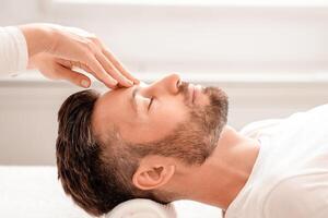 Unrecognizable female massage therapist rubbing relaxed man forehead, spa interior, side view. Handsome bearded man attending spa salon, getting lifting face massage. Male face treatment photo